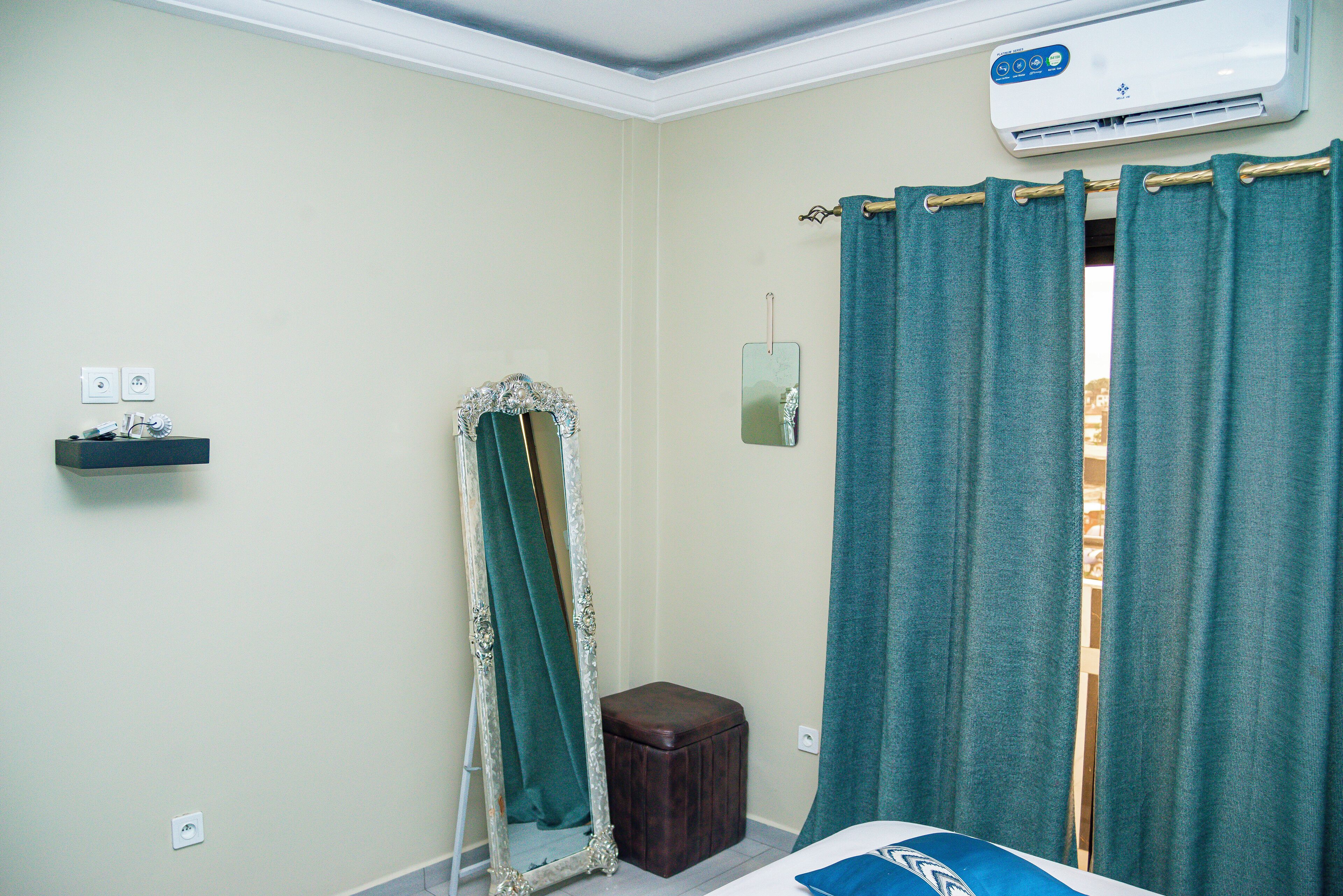 Habitació d'hotel amb un llit, un mirall, un petit tamboret, aire condicionat fixat a la paret i cortines color turquesa.