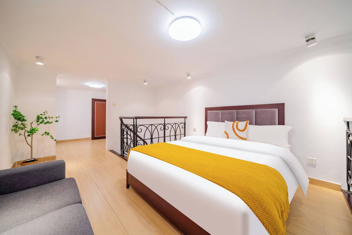 A modern hotel room with a large bed, a grey sofa, a potted plant, and a balcony with a railing.
