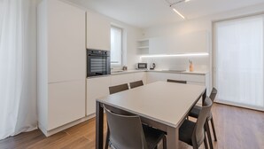 Et moderne køkken med hvide skabe, indbygget ovn, mikroovn og kaffemaskine. Der er et spisebord med fire stole.