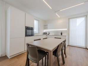 Cocina moderna con armarios blancos, horno empotrado, microondas y cafetera. Hay una mesa de comedor con sillas y un ventanal que proporciona luz natural.