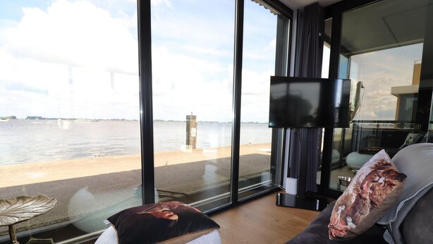 A modern living room with a large window overlooking a body of water, a flat-screen TV, and a small side table. - Houseboat With Sauna and Fantastic View (Offingawier)