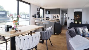 A modern kitchen with a central island, a dining area with a wooden table and chairs, and a living area with a sofa and cushions. - Houseboat With Sauna and Fantastic View (Offingawier)
