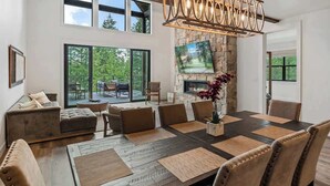 A spacious living area with a large wooden dining table, comfortable seating, a stone fireplace, and a large TV mounted on the wall. - Extraordinary Idaho Club Retreat w/ Hot Tub (Sandpoint)