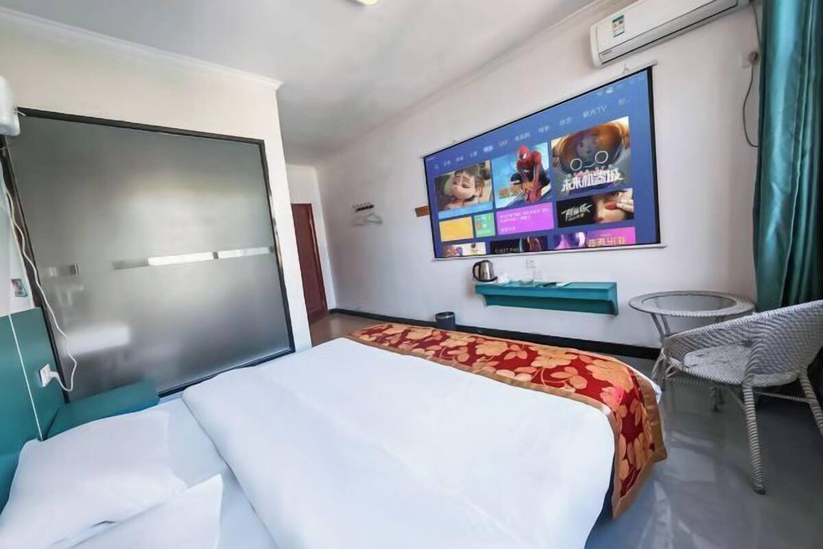 A hotel room with a bed, a television mounted on the wall displaying various channels, a small round table, and a wicker chair.