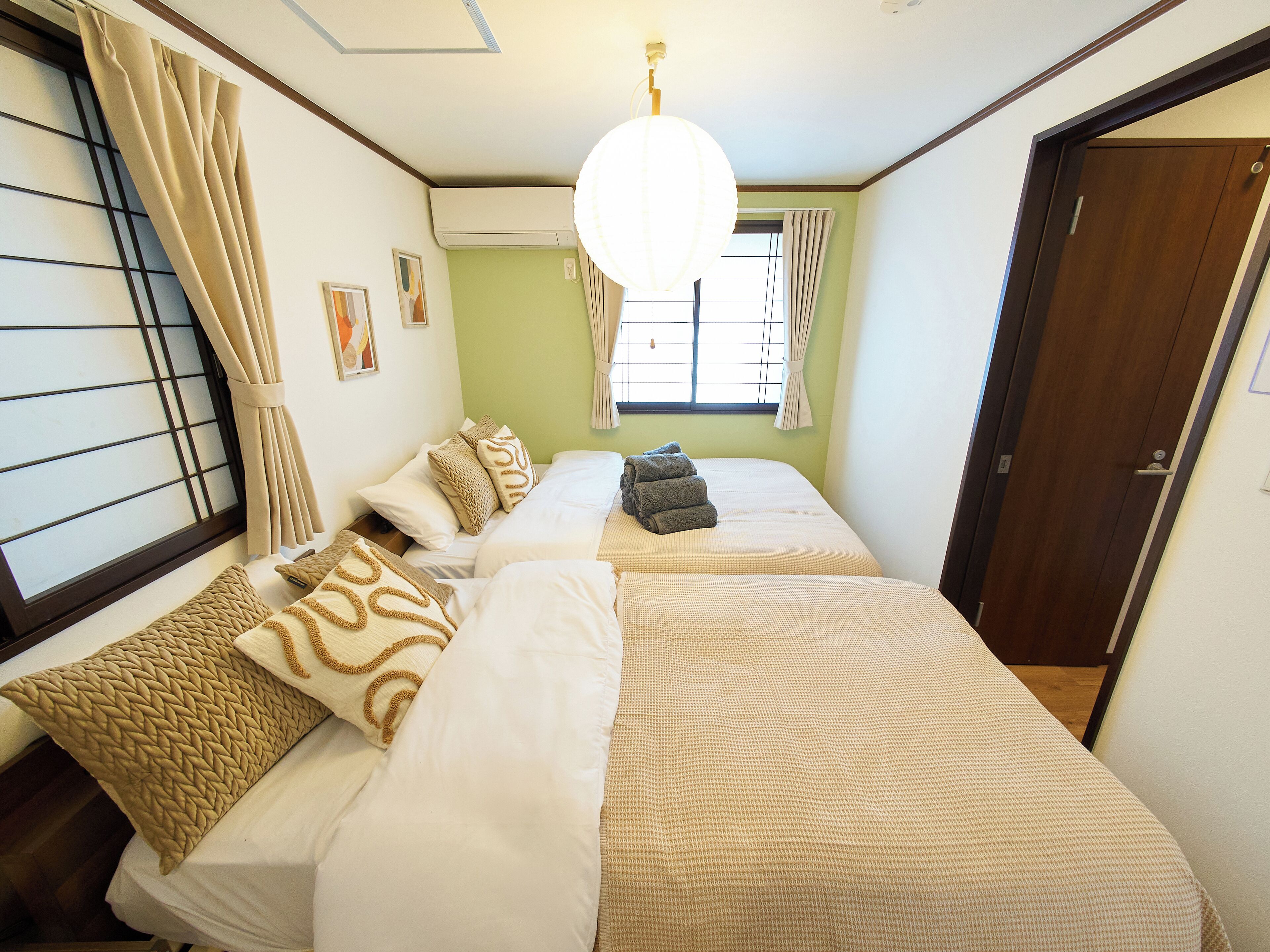 A room with two beds, beige and patterned bedding, a hanging light fixture, a window with curtains, and a door.