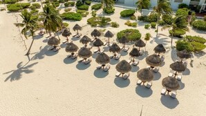An aerial view of a beach with rows of sun loungers and thatched umbrellas, surrounded by palm trees and landscaped greenery. - Exclusive Condo | 3BR | Beachfront & Top Amenities (Cancún)