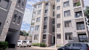 A multi-story residential building with balconies, a gated entrance, and parked cars.