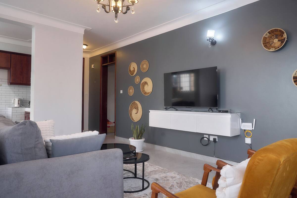 A modern living room with a grey sofa, a yellow armchair, a flat-screen TV mounted on a white shelf, decorative wall art, and a chandelier.