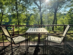 An outdoor deck with a table and two chairs, surrounded by trees and a view of the forest. - Charming 4-bedroom, riverfront cottage in Oswego (Oswego)