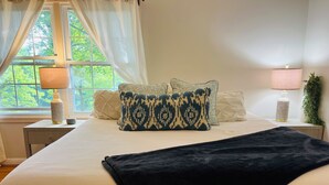 Een keurig opgemaakt bed met een donge deken, een raam met dunne gordijnen en een nachtkastje met een lamp en decoratieve objecten.
