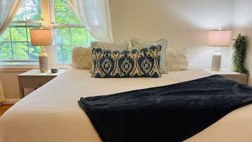 Un lit bien fait, recouvert d’un plaid sombre, une fenêtre ornée de rideaux légers et une table de chevet avec une lampe et quelques objets décoratifs.