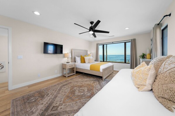 A modern hotel room with a large bed, a ceiling fan, a TV, and a view of the ocean.