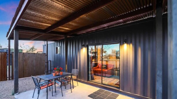 Un patio couvert avec une table et des chaises, une porte-fenêtre coulissante donnant sur un espace extérieur aménagé, et une structure de toit en métal.