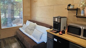 A compact room with a wooden floor, a brown sofa with white cushions, a coffee maker, a microwave, and a small shelf with a vase. - Tiny Home Retreat with hot tub (Lake Villa)