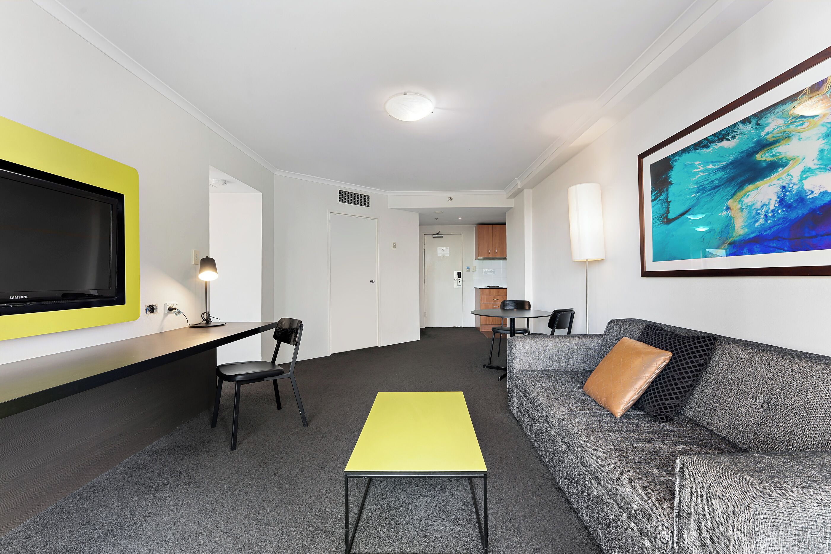 Une chambre d’hôtel moderne équipée d’un téléviseur à écran plat, d’une table jaune, d’un canapé gris avec un coussin marron et d’un grand tableau abstrait accroché au mur.