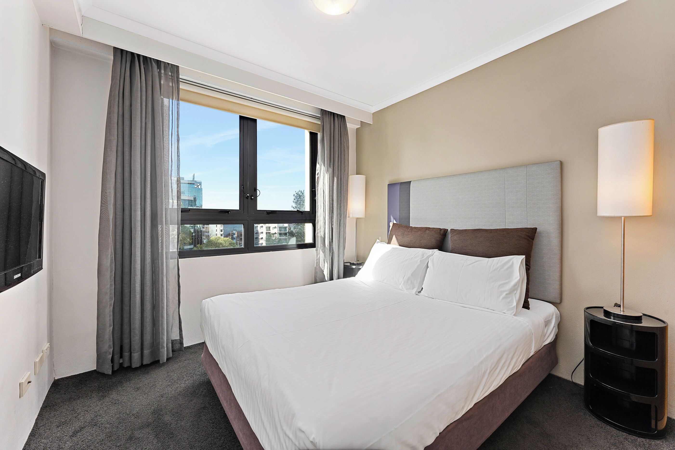 A hotel room with a large bed, a TV, a window with curtains, and a lamp.