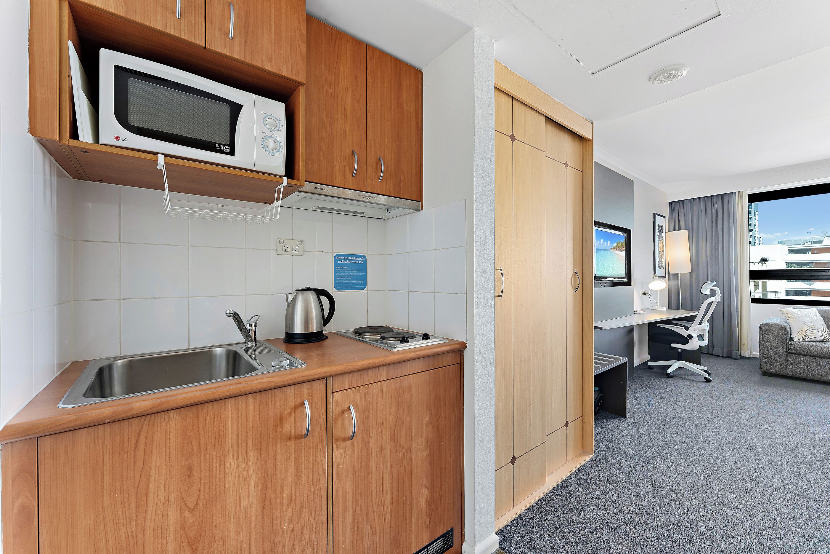 A compact kitchen with wooden cabinets, a microwave, a kettle, and a sink. There is a desk with a chair and a window with a view of the city.