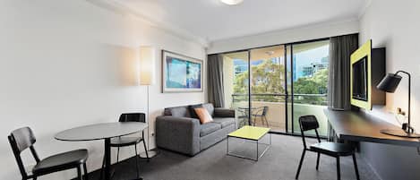 Una habitación de hotel moderna con un sofá gris, una pequeña mesa redonda, dos sillas, una mesa de centro amarilla, un escritorio con lámpara y un ventanal con vistas a edificios y árboles.