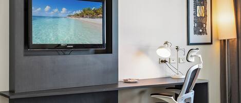 A hotel room with a flat-screen TV displaying a beach scene, a desk with a chair, a framed artwork, and a bedside lamp.