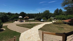 Uma trilha de pedra leva a uma área de piscina com um gazebo e um pequeno edifício, tudo cercado por árvores e um céu azul límpido.