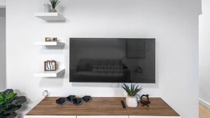 A modern living room with a large flat-screen TV mounted on the wall, a wooden coffee table, and white shelves with decorative items. - Centric Modern Boho Apt For 6 @ San Juan (San Juan)