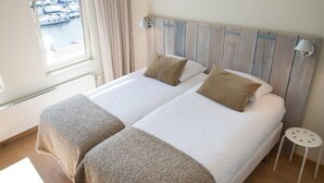 A double bed with two pillows, a wooden headboard, a small bedside table, and a view of boats through a window. - Apartment, Near the Beach and the sea (The Hague)