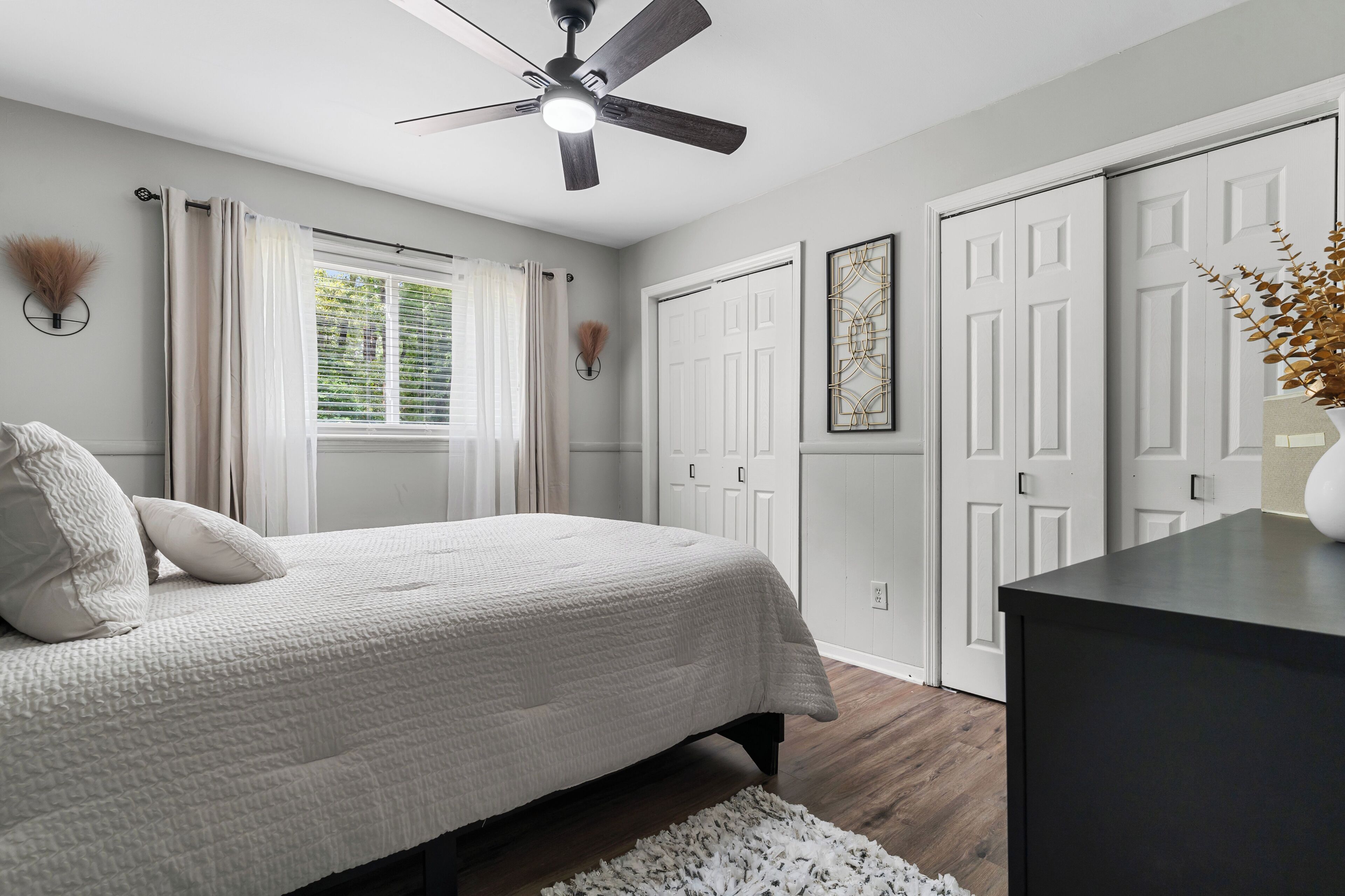 A bedroom with a bed, ceiling fan, white curtains, and a window.