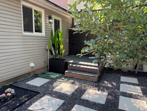 A residential outdoor area with a wooden deck, a black planter with large green leaves, and stepping stones on gravel.