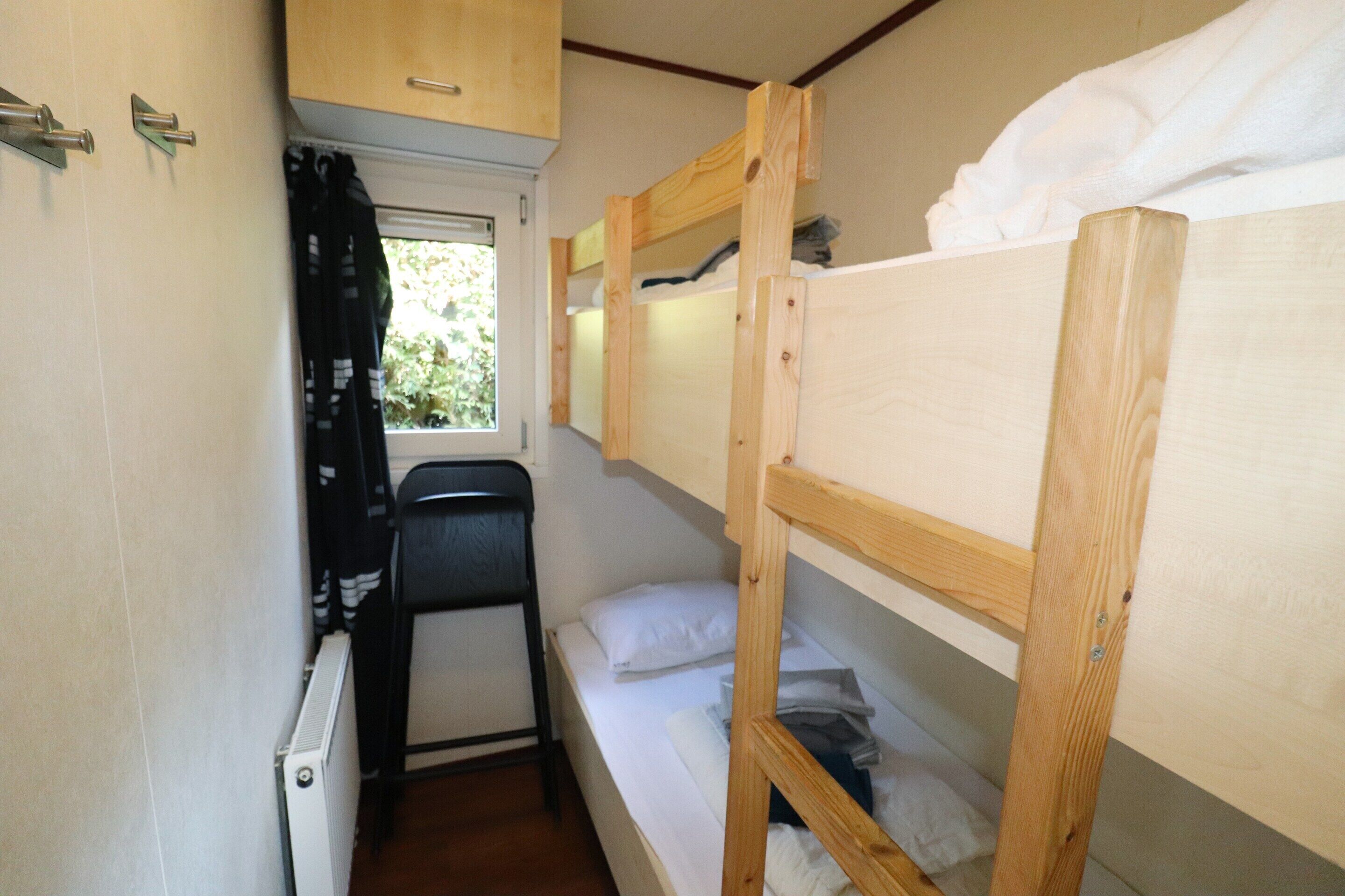 A small, compact room with bunk beds, a radiator, a window with curtains, and a chair.