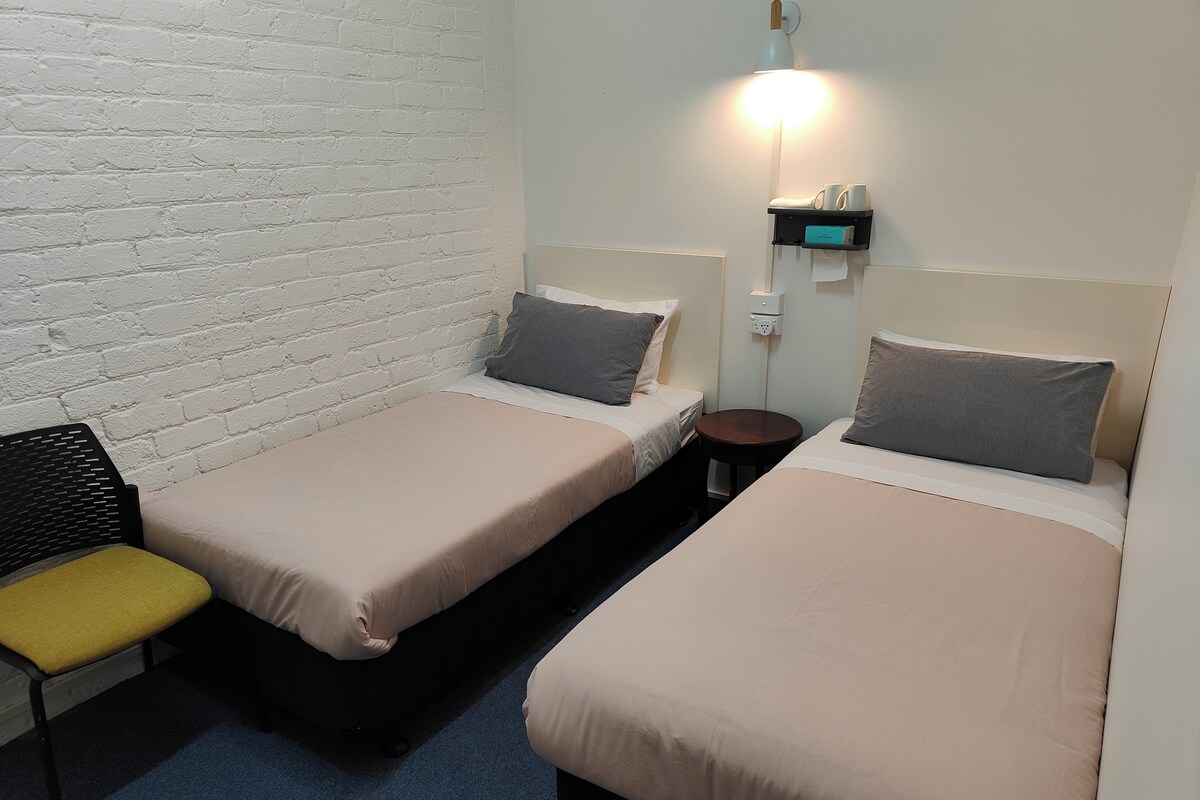 A small, clean room with two single beds, a chair, a small table, and a wall-mounted lamp.
