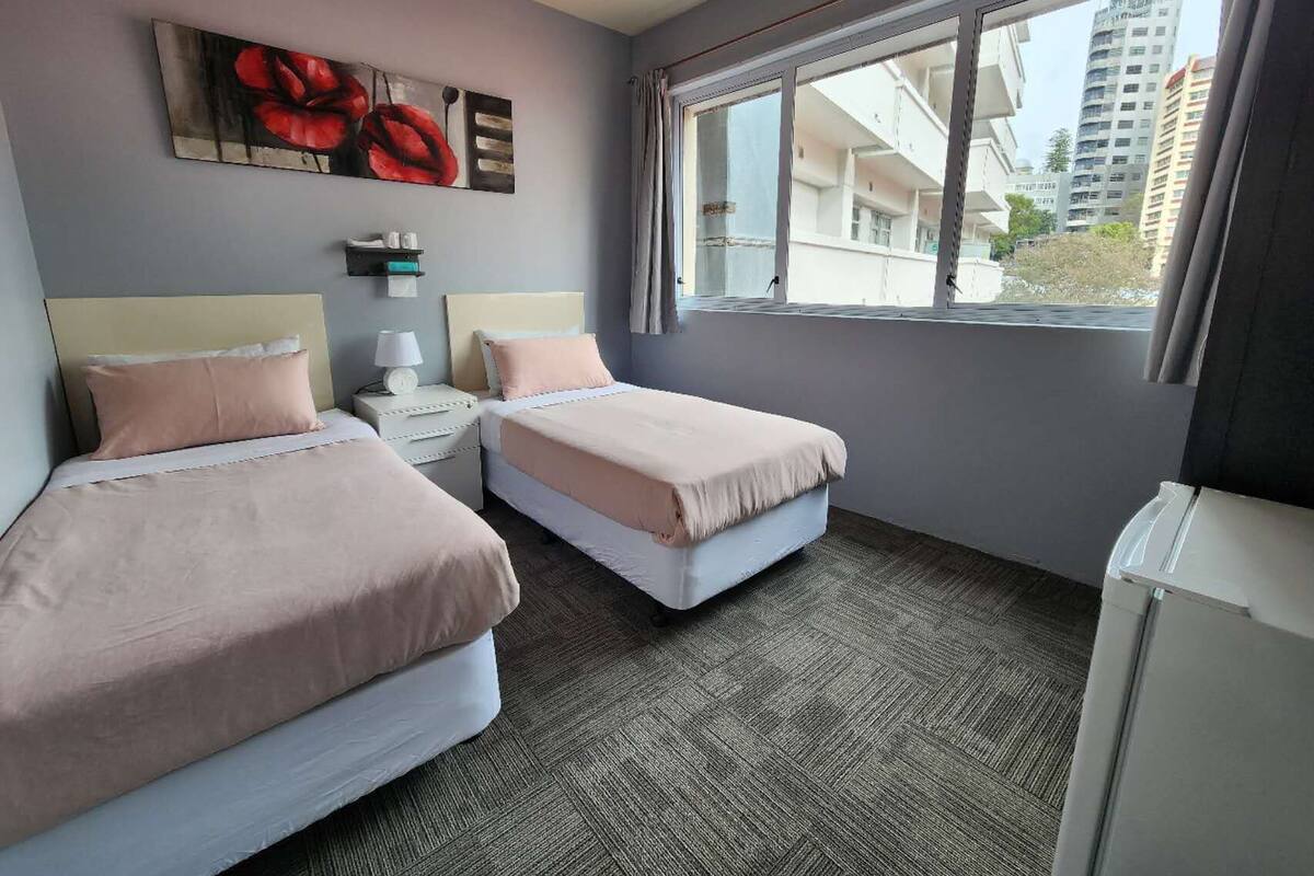 Une chambre d’hôtel avec deux lits, une table de chevet avec une lampe, un tableau au mur et une fenêtre donnant sur des immeubles.