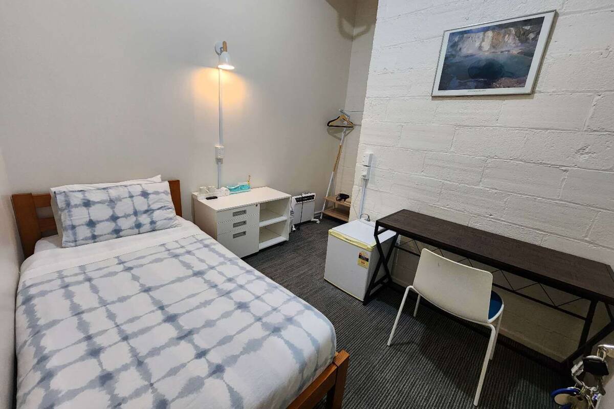 Une petite pièce bien éclairée, avec un lit, un bureau, une chaise et un tableau accroché au mur.
