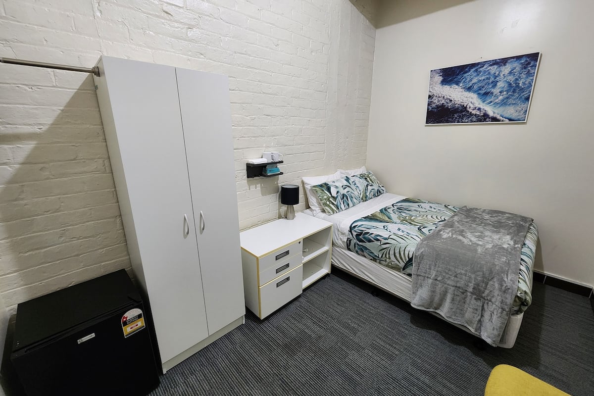 Une chambre à coucher de taille modeste, comprenant un lit, une armoire blanche, un bureau avec une lampe et une petite poubelle.