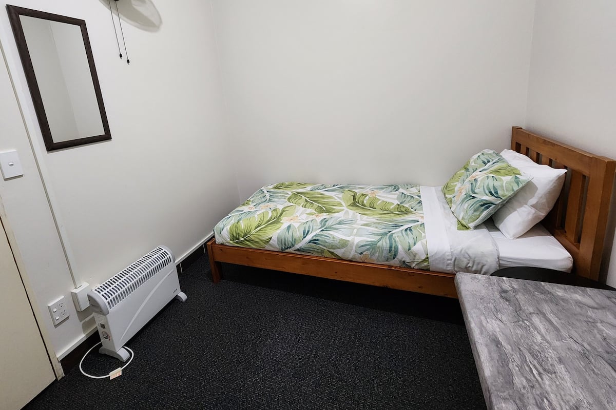 Une petite chambre avec un lit simple, une tête de lit en bois, une couvre-lit à motifs floraux, une table de chevet et un radiateur.