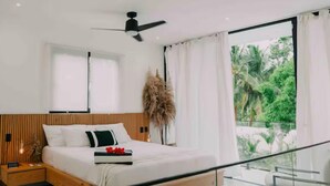 A modern bedroom with a wooden headboard, a bed with white bedding, a bedside table with a lamp, and a view of greenery through large windows. - Brand new 3-bedroom Villa & 1-bed bungalow  Las Terrenas with AC, WiFi (Las Terrenas)