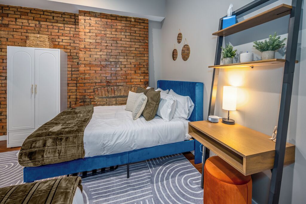 A bedroom with a blue bed, white wardrobe, brick wall, and a wooden desk with a lamp.