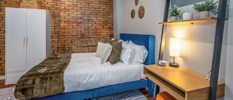 A bedroom with a blue bed, white wardrobe, brick wall, and a wooden desk with a lamp.