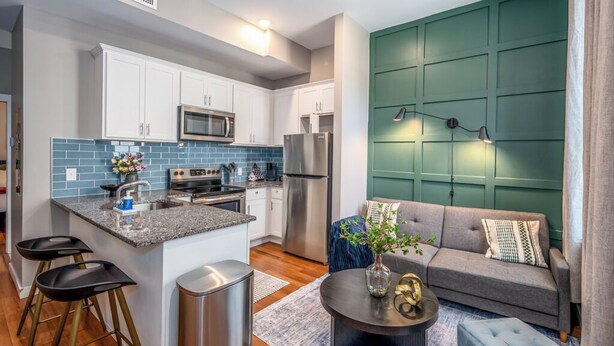 A modern kitchen with white cabinets, a granite countertop, and stainless steel appliances. There is a grey sofa and a round coffee table with a vase and a decorative bowl.