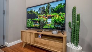 A flat-screen TV on a wooden TV stand with a remote control and a small sign. A large cactus is in a white pot beside the TV.