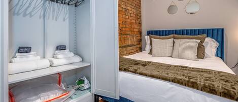A hotel room with a neatly organized closet, a bed with a blue headboard, and a brick wall.