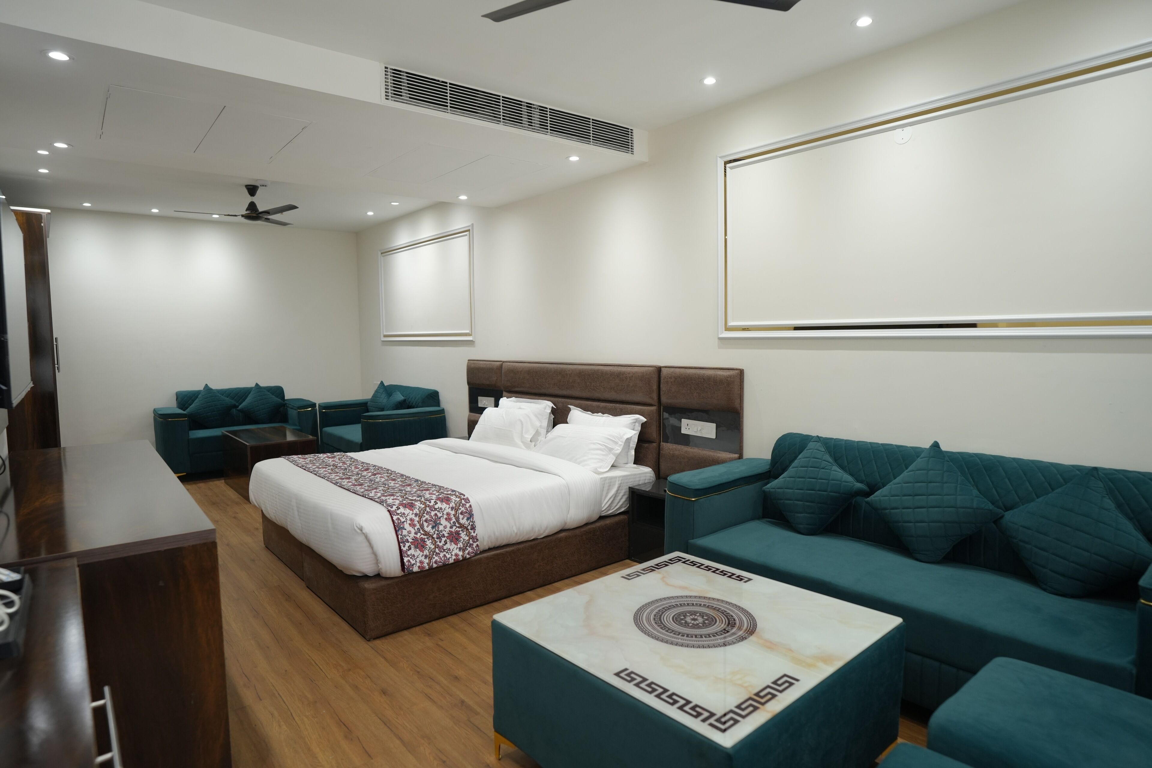 A modern hotel room with a large bed, two green sofas, a wooden desk, and a marble coffee table.