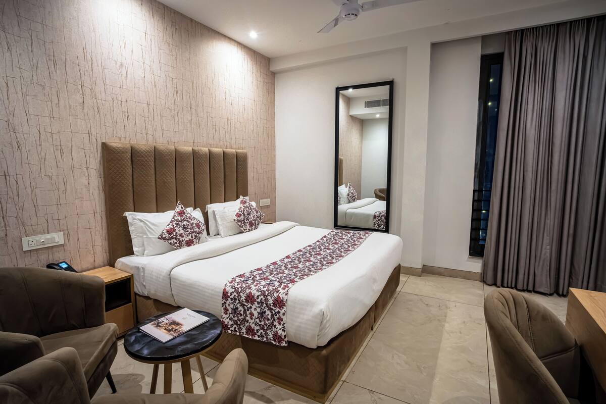 A hotel room with a large bed, a bedside table, a chair, a small table, and a mirror.