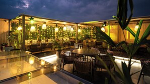 An outdoor dining area with round tables and wicker chairs, surrounded by potted plants and illuminated by hanging lanterns. - ACR Luxury Hotel (Mohali)