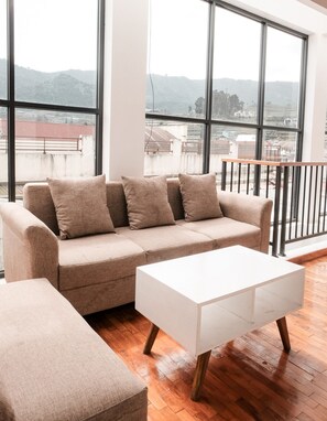 Un soggiorno moderno con un divano color beige, un tavolino da caffè bianco e ampie finestre che offrono una vista sulla città e sulle colline.