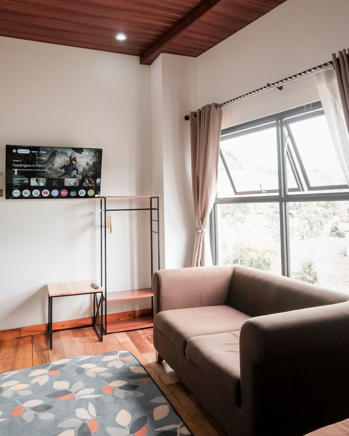 A modern living room with a sofa, a small table, a TV mounted on the wall, and a window with curtains.