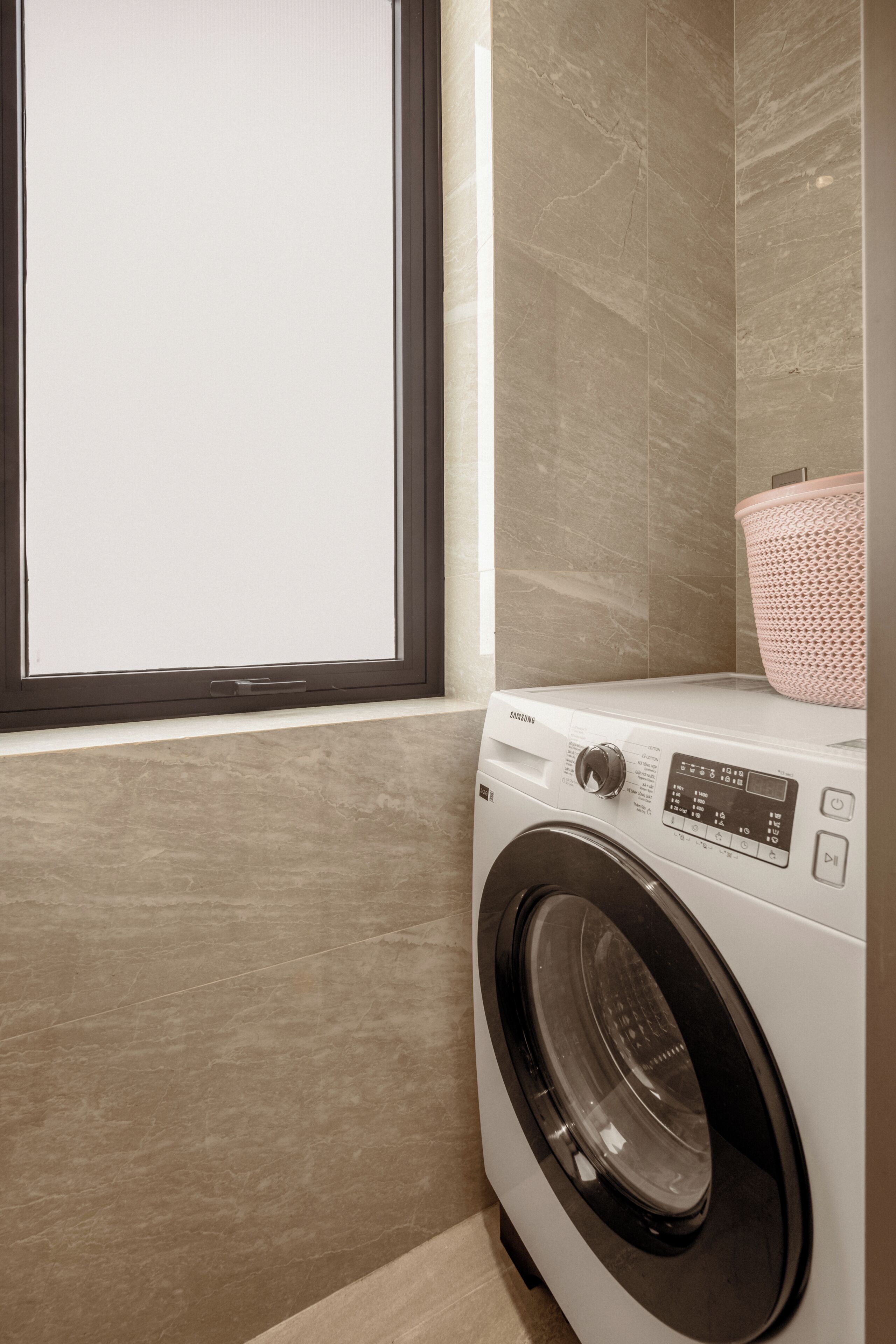 A front view of a front-loading washing machine in a laundry room with a window and a tiled wall.
