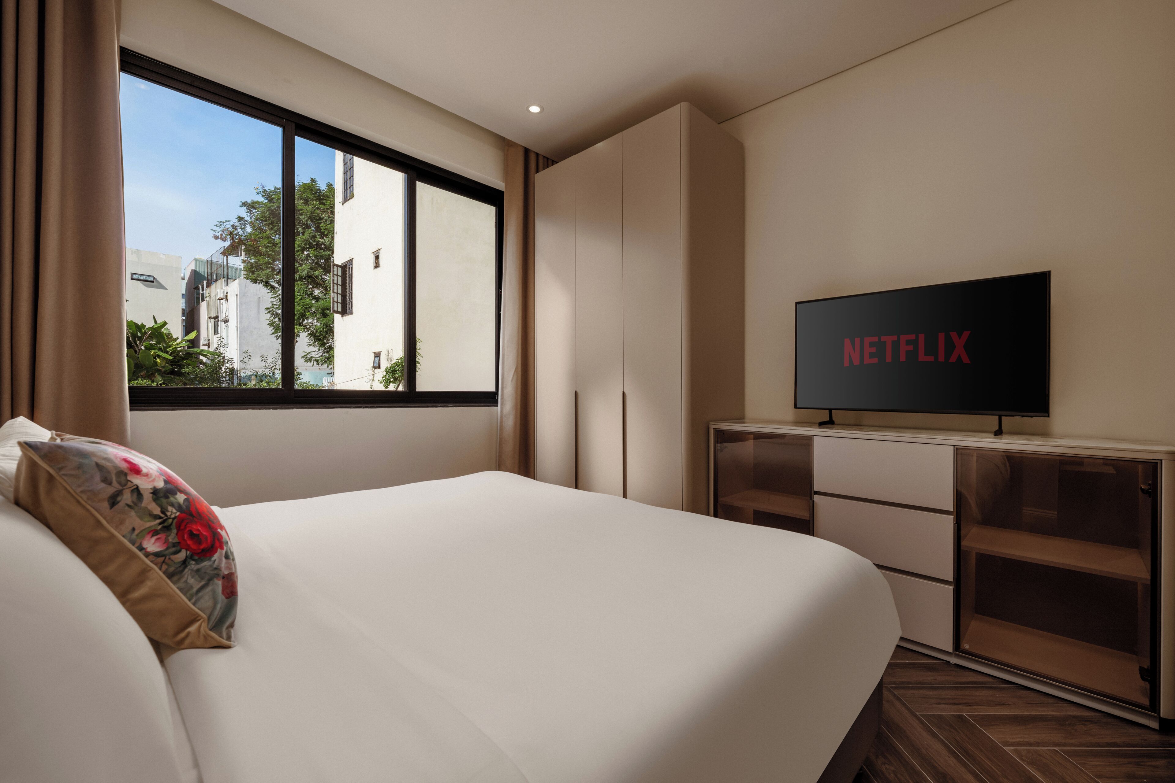 A hotel room with a bed, a television on a stand, and a window with a view of buildings.