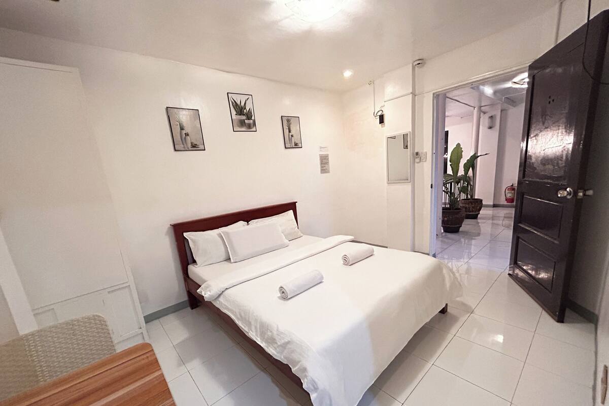 A hotel room with a large bed, a wooden bench, a small table, and a door leading to a hallway with plants.