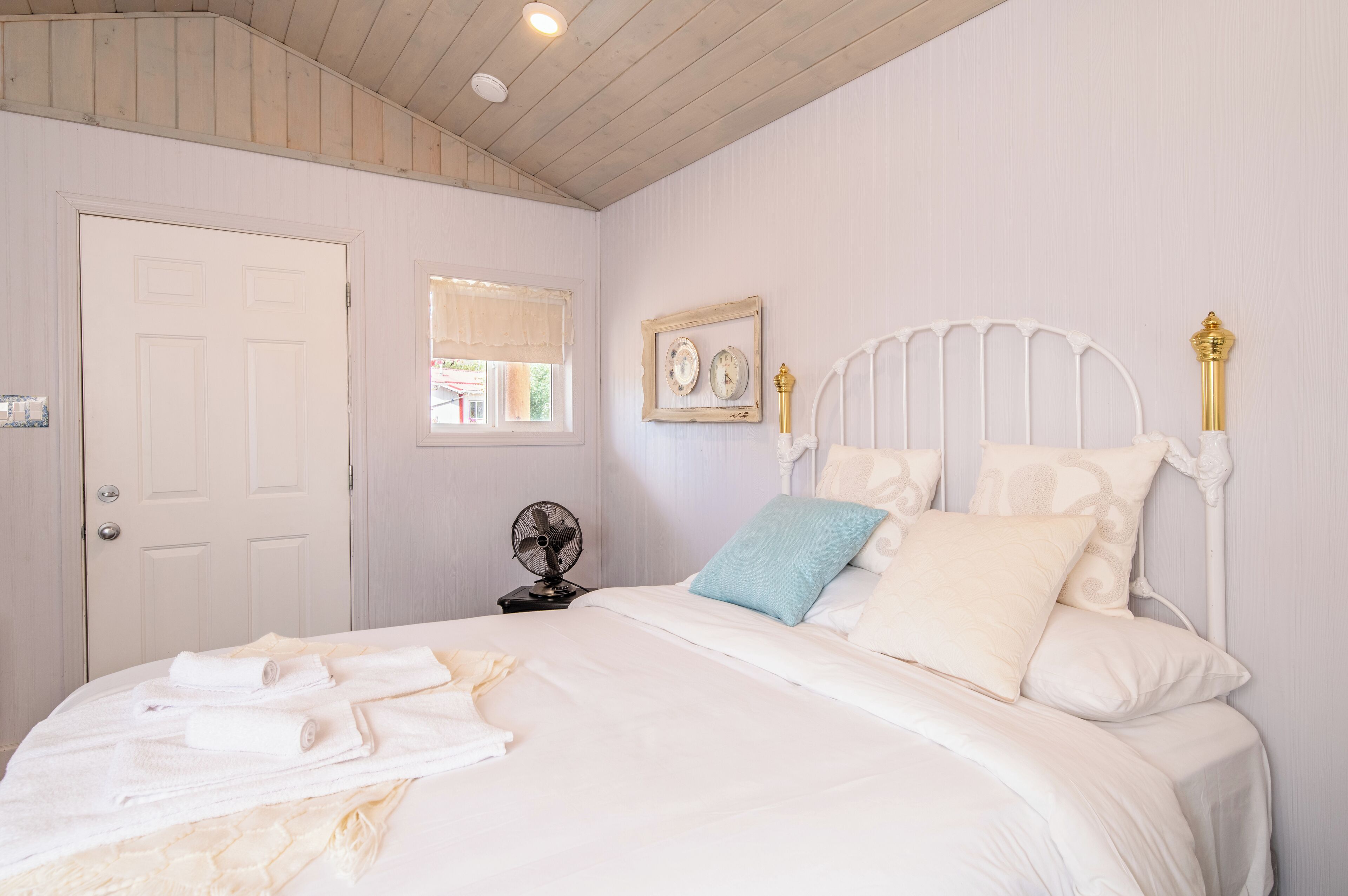 A bedroom with a bed, white walls, a wooden ceiling, a door, a window with a curtain, a bedside table with a fan, and a framed picture on the wall.