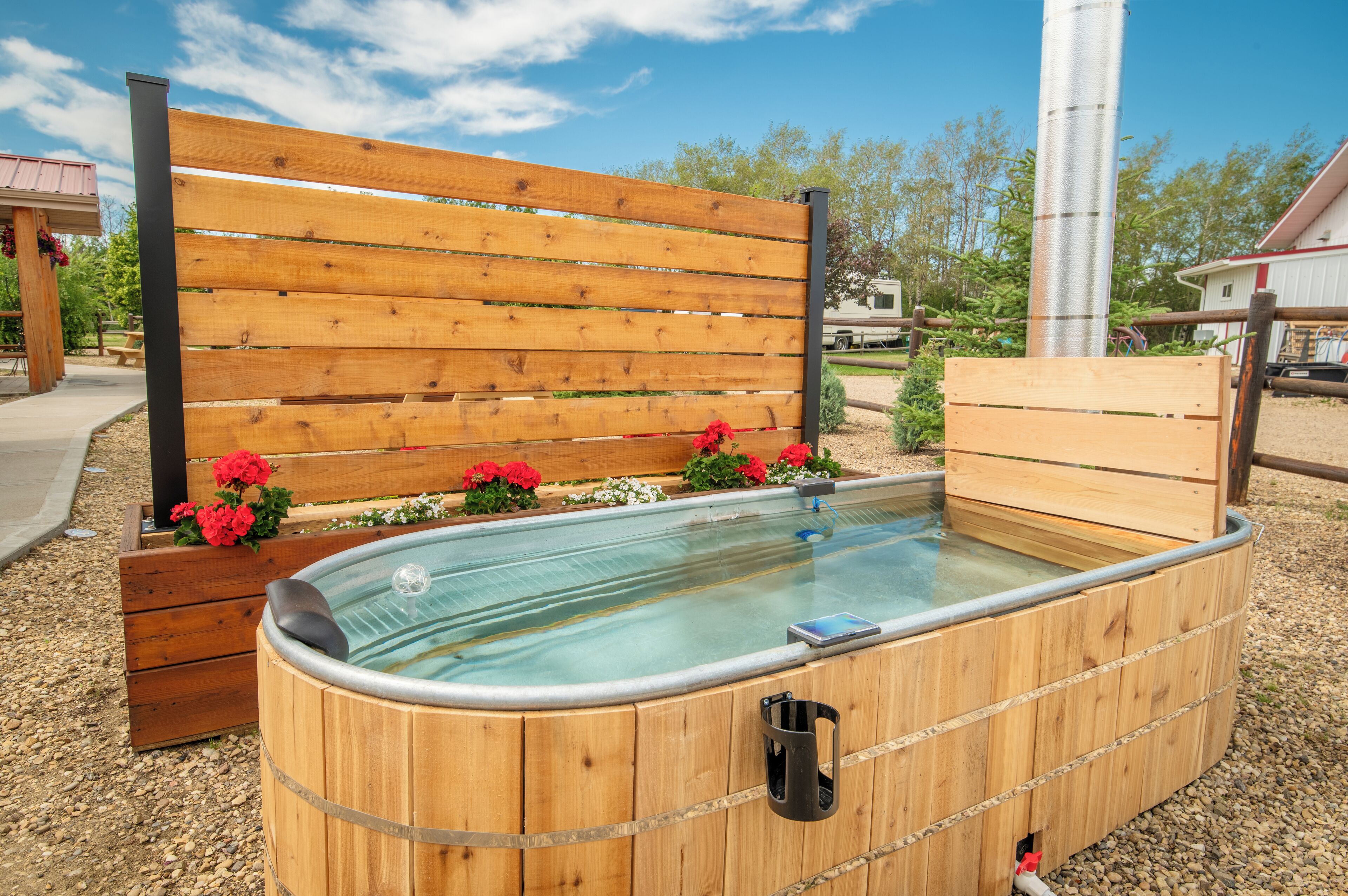 A wooden hot tub with a bench and flowers, surrounded by gravel and trees.